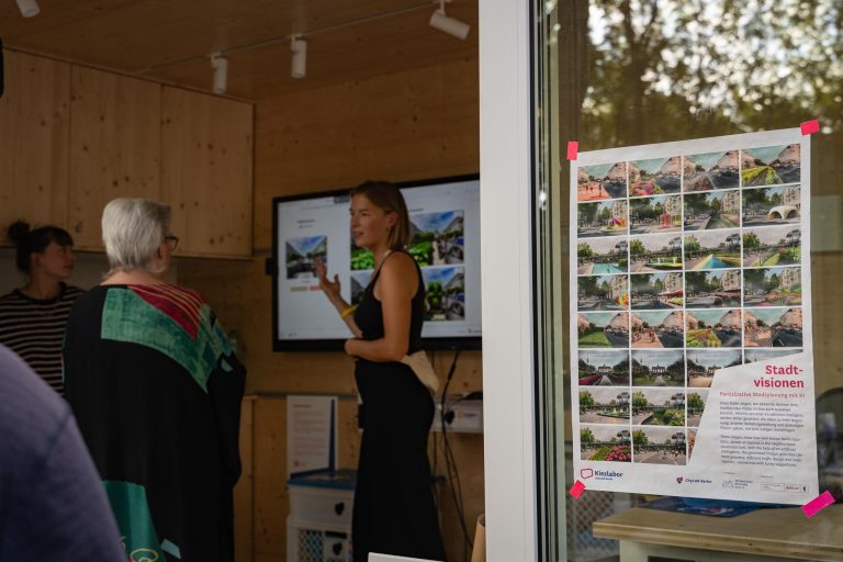 Eine Frau steht vor einem Bildschirm und präsentiert etwas. Man erkennt, dass es um Stadtvisionen und visualisierten Utopien geht. Man kann ZuhörerInnen erkennen.