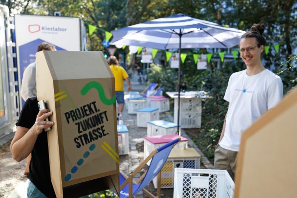 Eine Person schaut durch ein Periskop, durch das man die Straße durch Kinderaugen sehen kann. Auf dem Modell steht "Projekt Zukunft Straße".